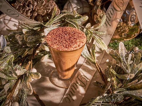 Tiramisu eggnog cocktail topped with chocolate shavings on a table with festive decor, pinecones and greenery and ribbon with the word Dante